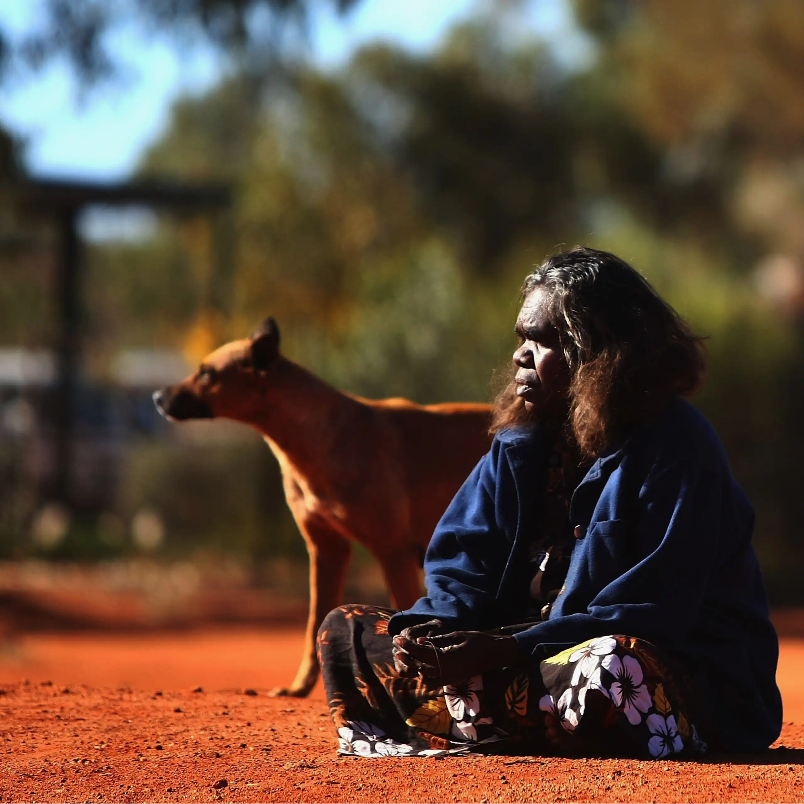 An Indigenous woman sits on red dirt with a dog