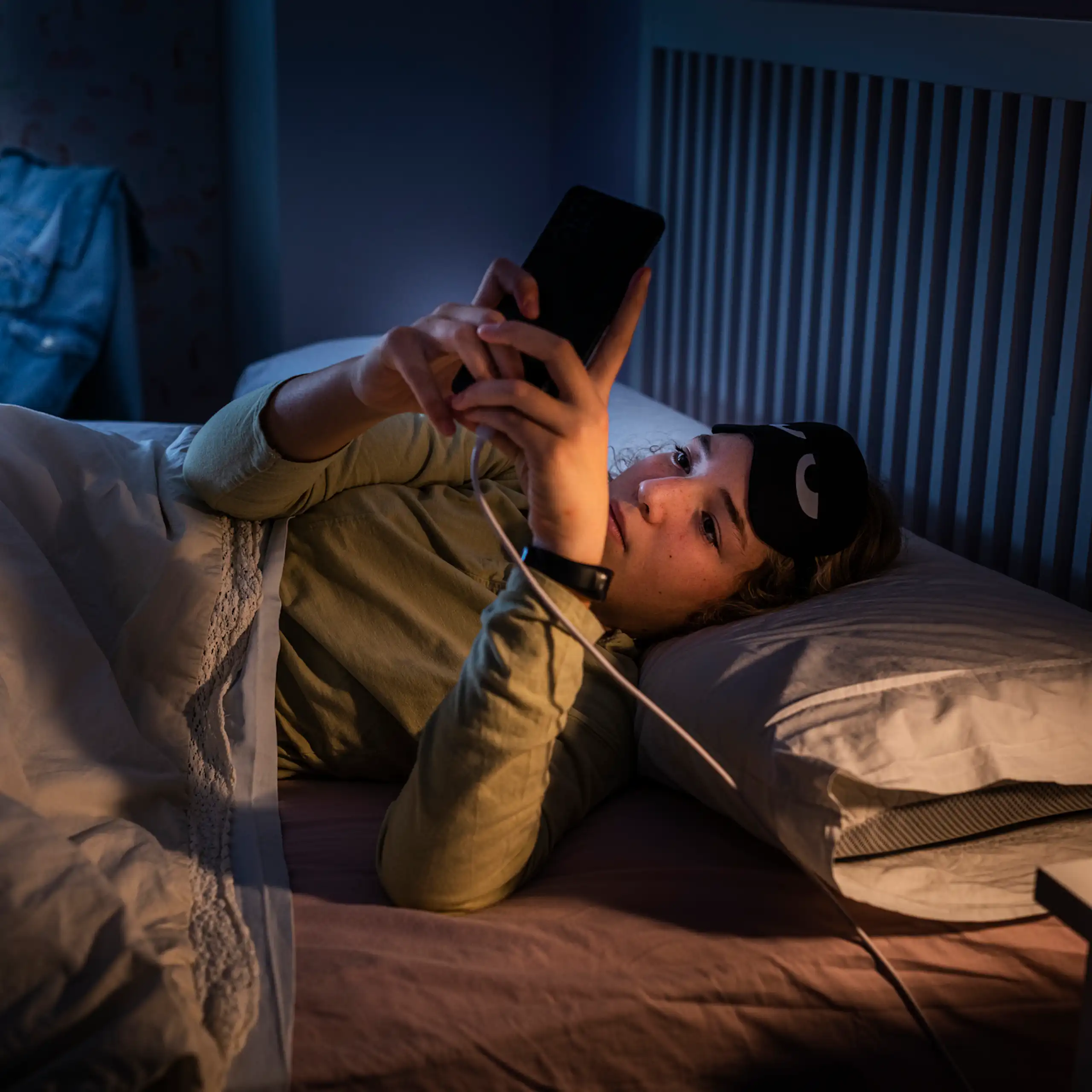 A woman at night looking at her phone that's plugged in by her bed.