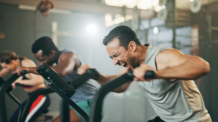 Two men push themselves to their limits while exercising on a cardio machine in the gym.