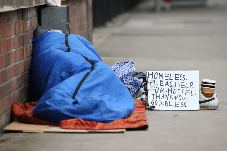 A rough sleeper next to a sign asking for help.