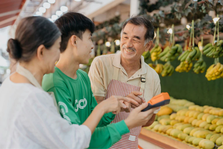 A couple considers buying fruit at a grocery store, with bananas in the background, while a smiling manager offers help.