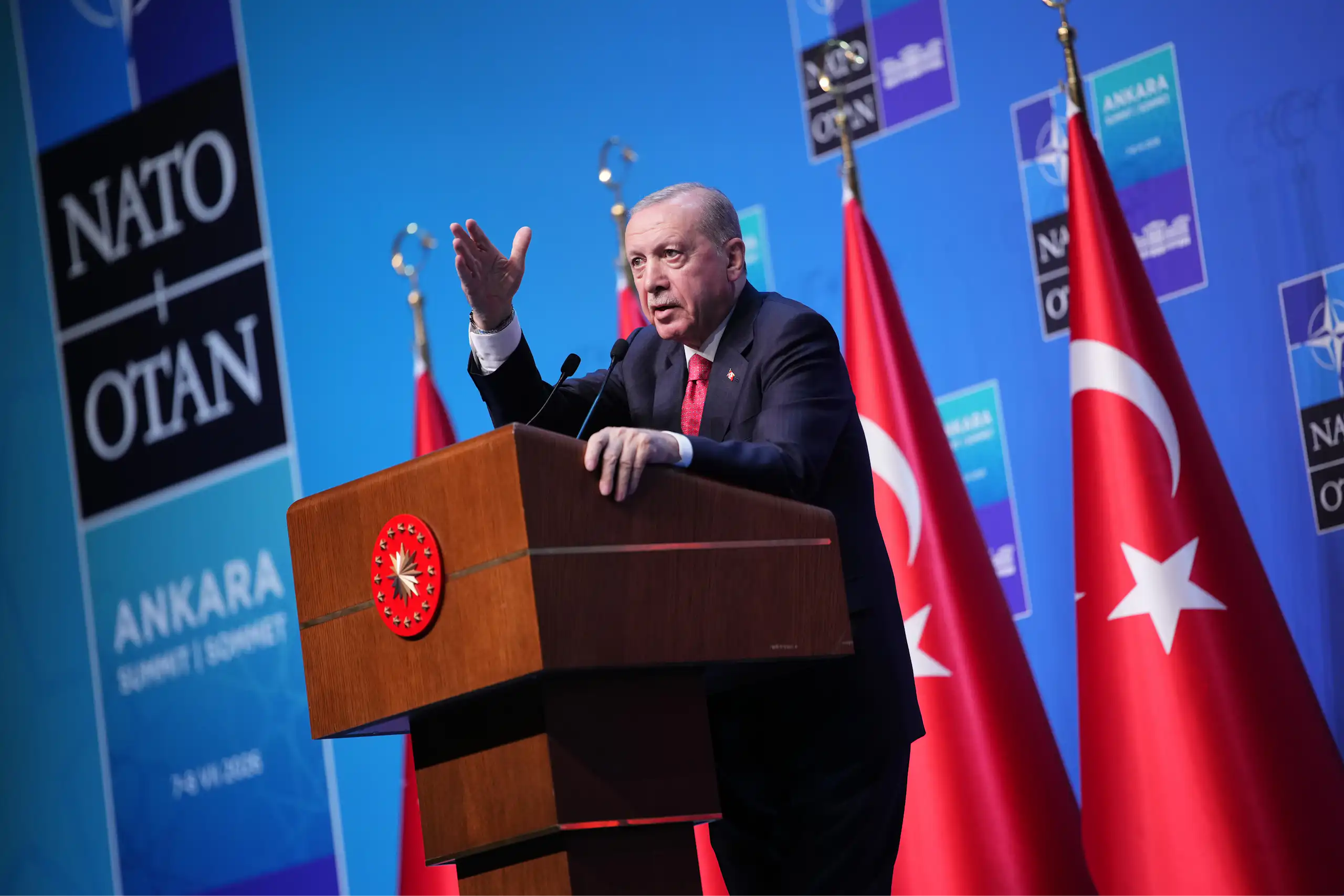 A man speaks from a podium next to red flags. A NATO banner is behind him.
