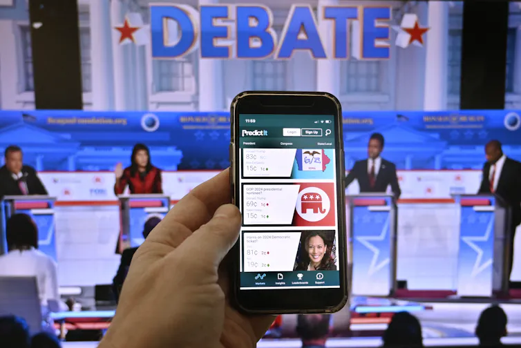 A smartphone is seen in the foreground as four people stand in front of podiums on a stage in the background.