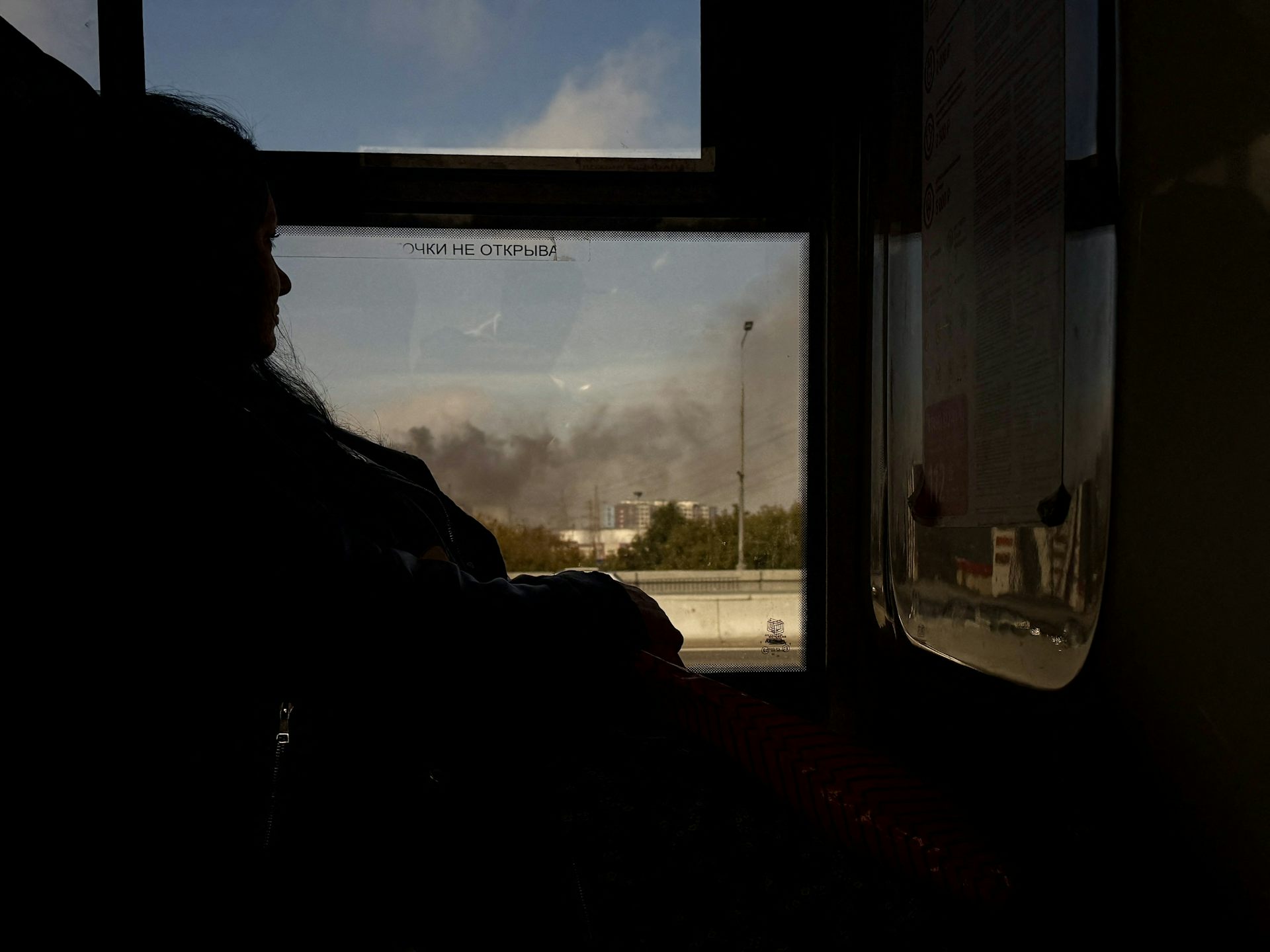 A woman looks out a window at smoke billowing from a building in the distance.