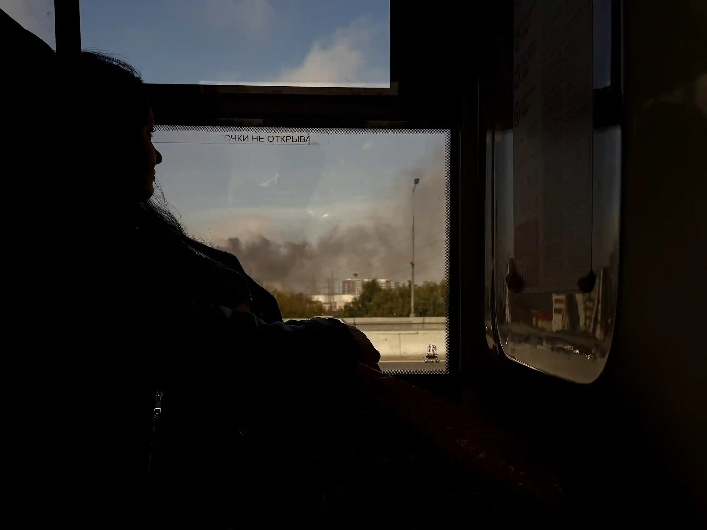 A woman looks out a window at smoke billowing from a building in the distance.