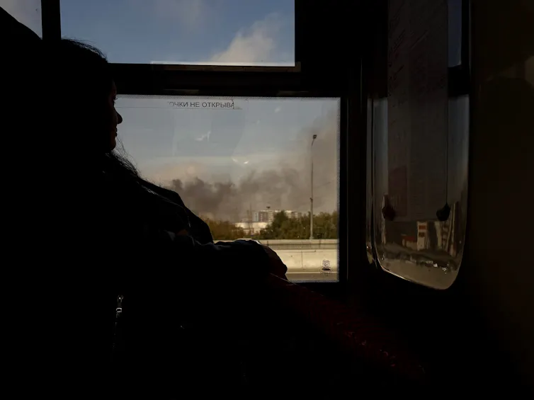 A woman looks out a window at smoke billowing from a building in the distance.