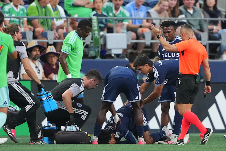 One man on the ground on a soccer pitch with his head in his hands, while other players, a referee and medical staff gather around, wearing bright green, orange, blue and black shirts.