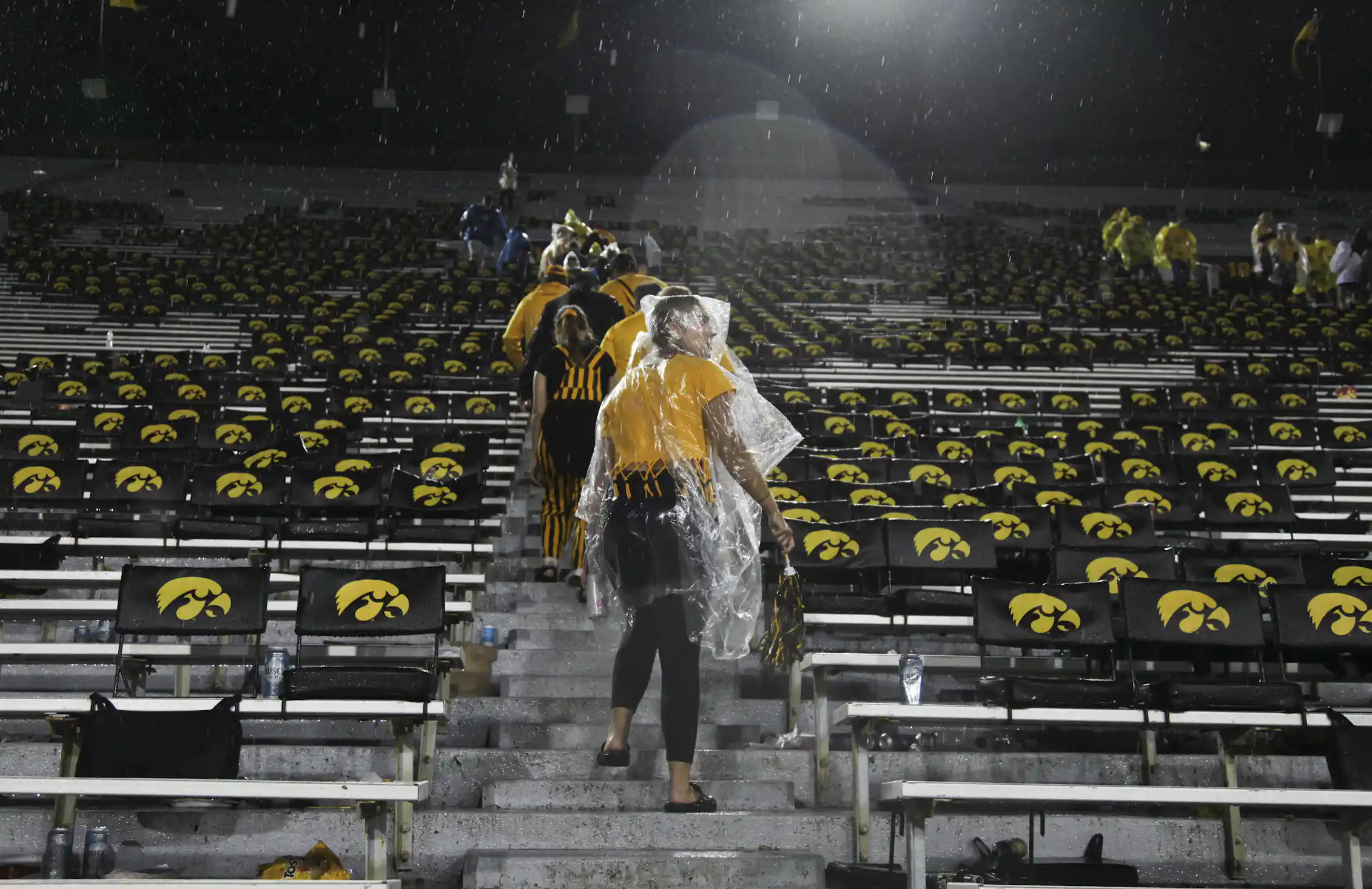 People with raincoats over their heads walk up stairs with Iowa Hawkeyes symbols on the seats around them.