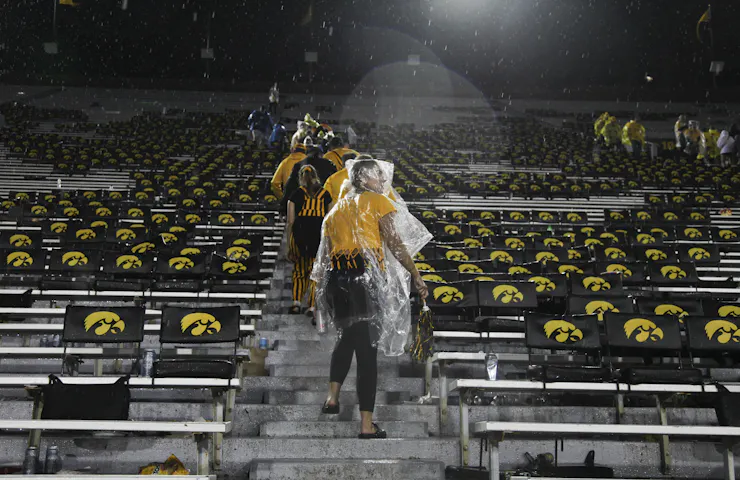 People with raincoats over their heads walk up stairs with Iowa Hawkeyes symbols on the seats around them.