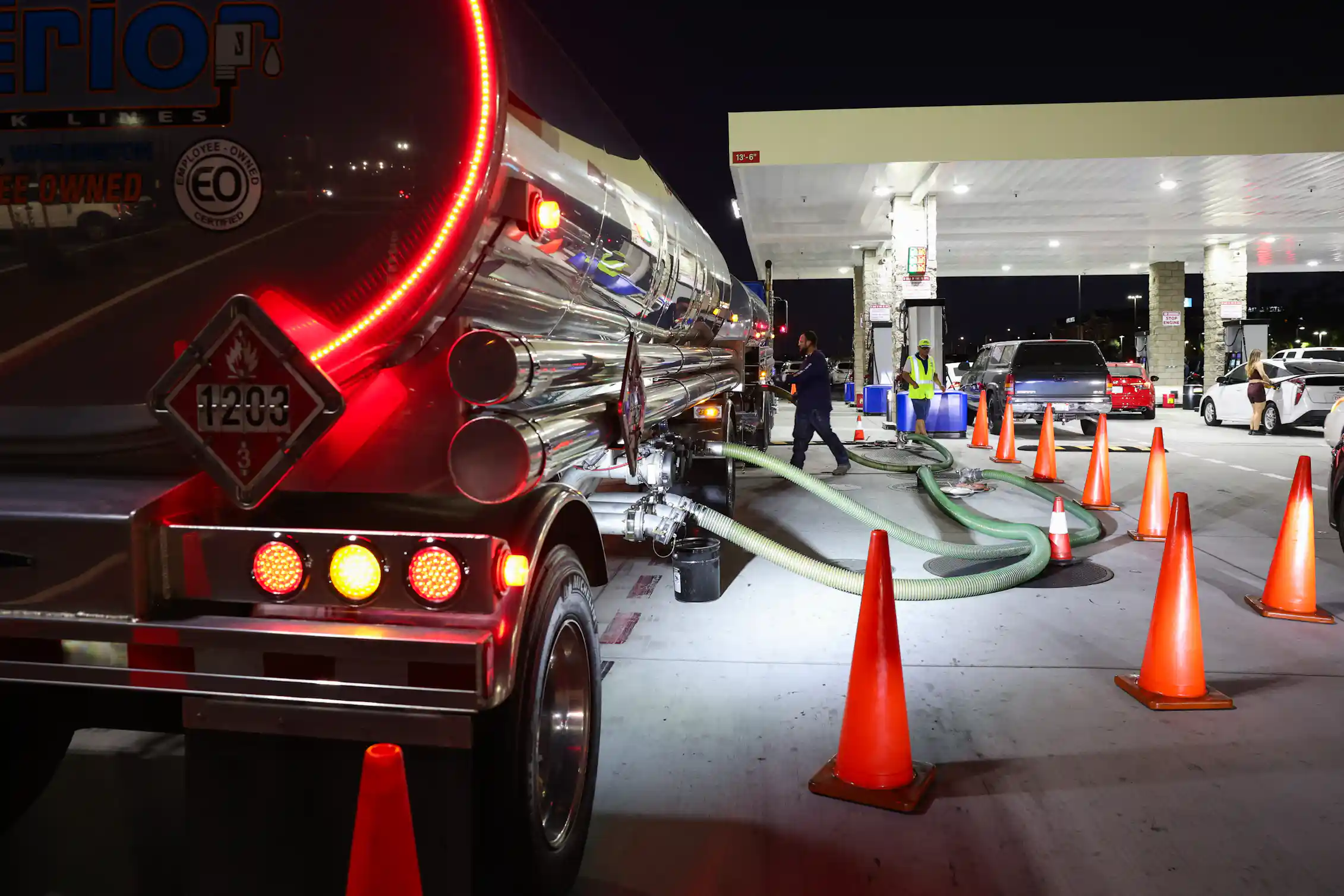 A large tanker truck sits next to a gas station, with tubes stretching from the tank into the ground.