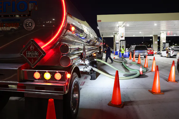 A large tanker truck sits next to a gas station, with tubes stretching from the tank into the ground.