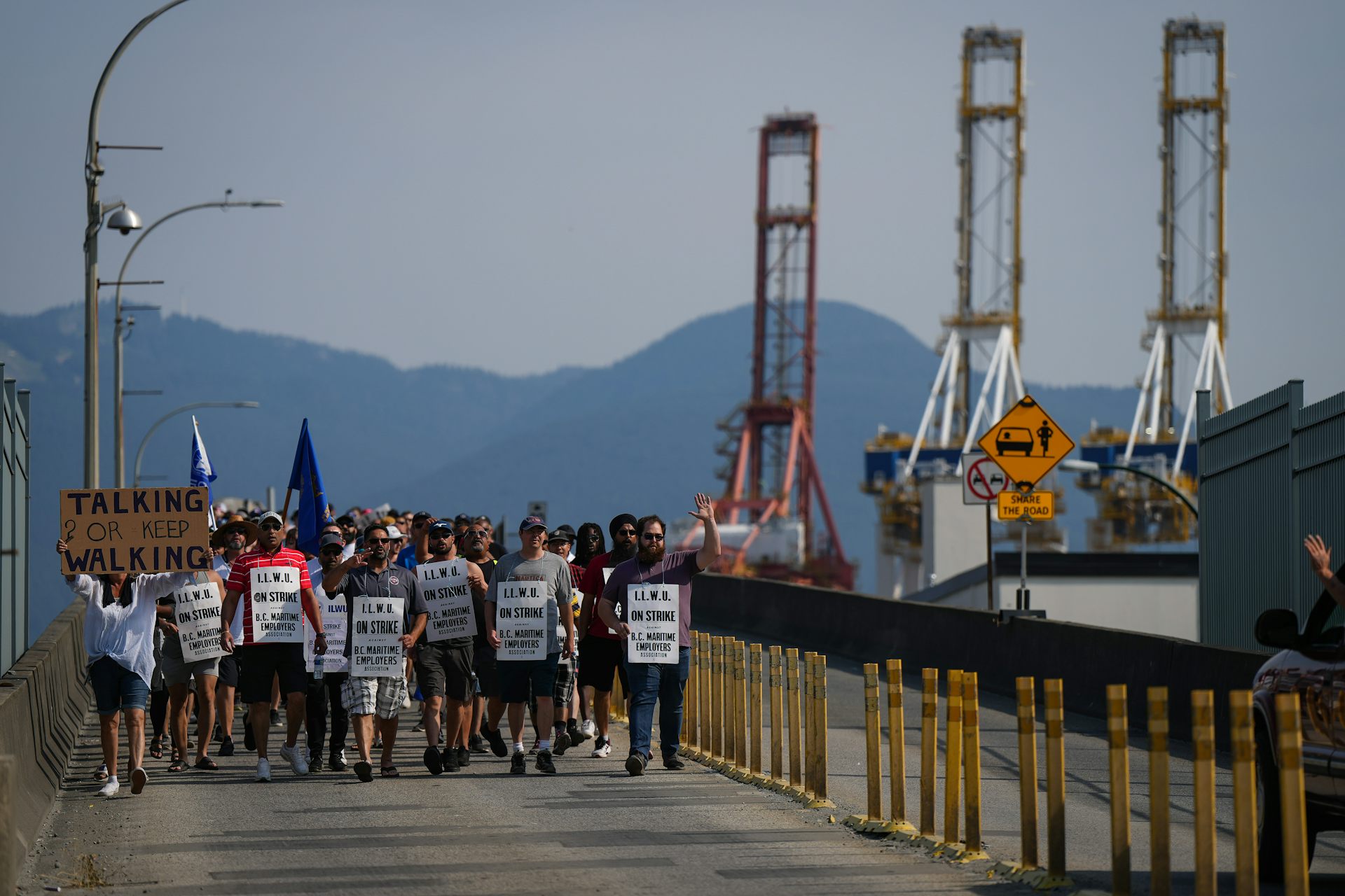 People carrying picket signs walk past a port where cranes load and unload cargo