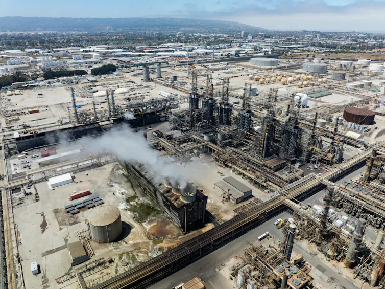 An aerial view shows an industrial site with tanks and pipes and smokestacks.