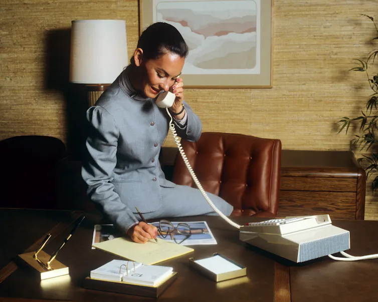 A woman in a blue suit sits on a desk and talks on the phone.