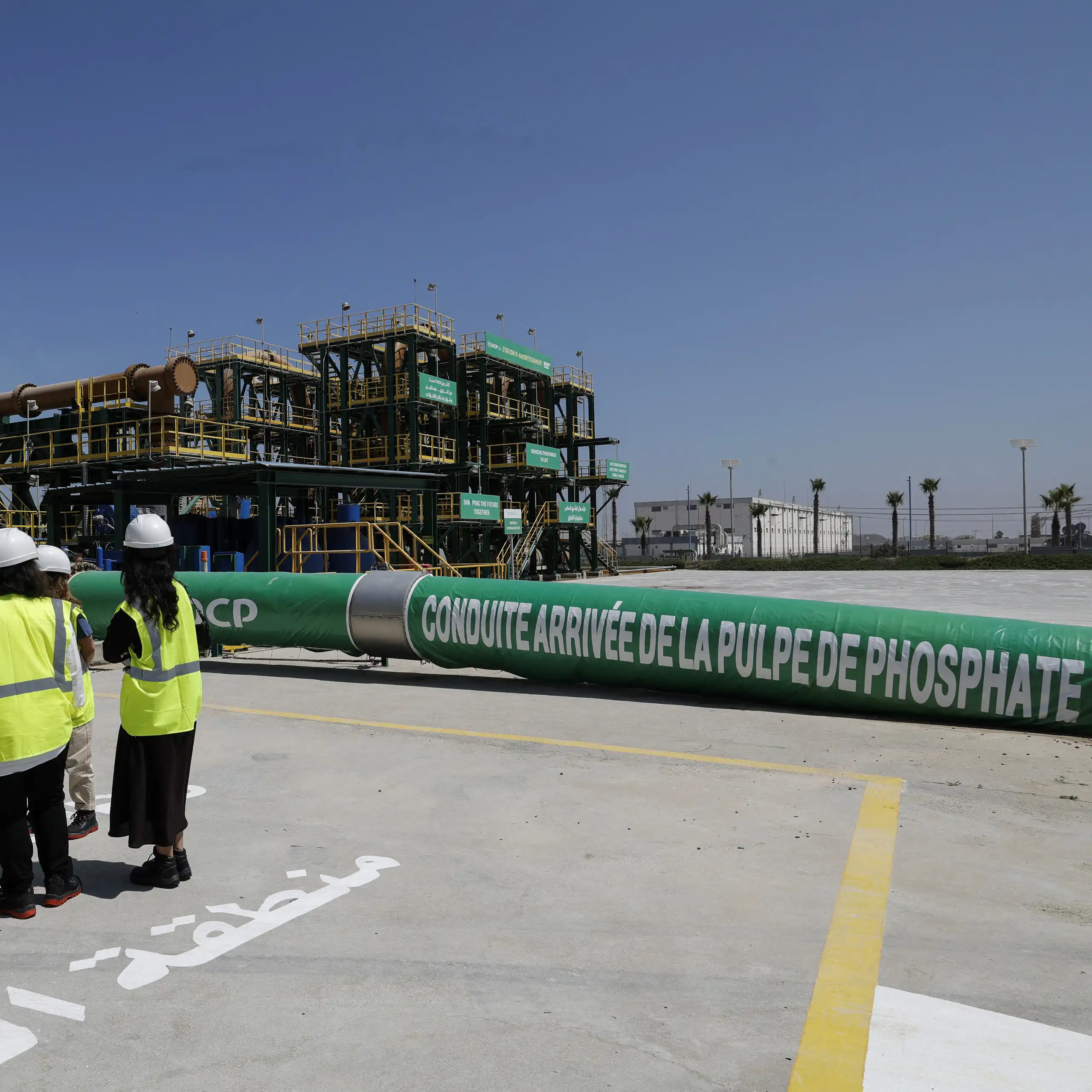 People in hard hats and yellow reflective vests stand next to a long green pipeline that says phosphate on it