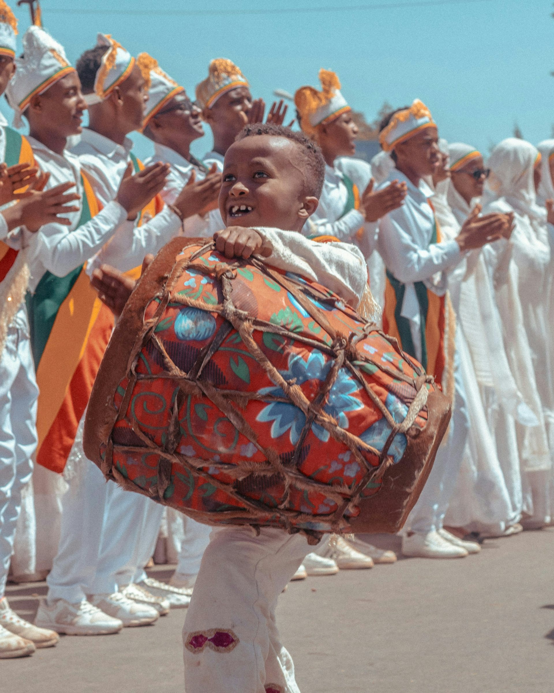 A boy playing a drum while standing in front of a row of men dressed in white robes