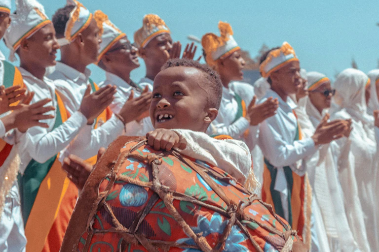 A boy playing a drum while standing in front of a row of men dressed in white robes