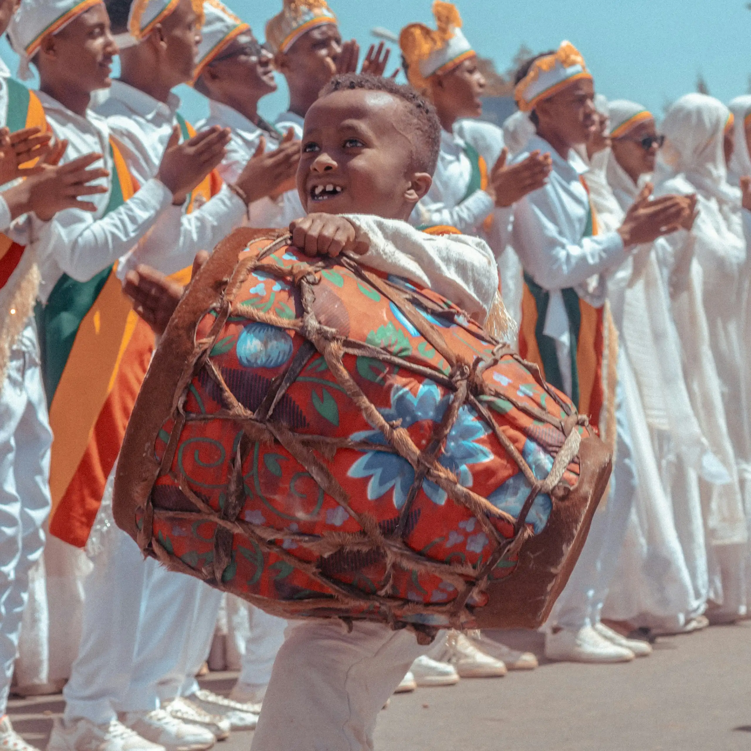 A boy playing a drum while standing in front of a row of men dressed in white robes