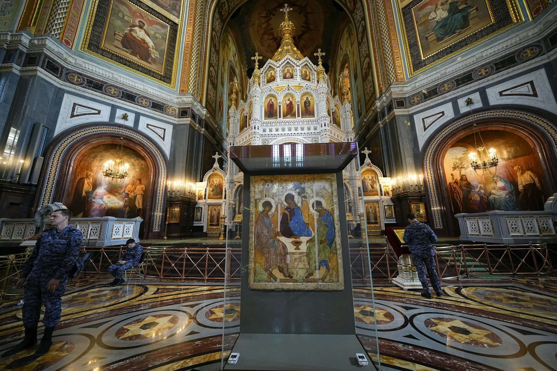 Men in military fatigue stand next to a religious icon of three angel figures behind a class case inside an ornate church with gold decor.