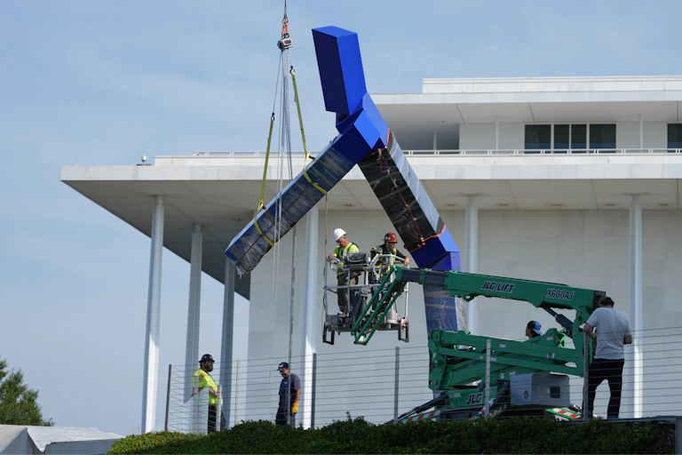A crane with workers seen lifting a blue sculpture that seems to be in a large Y shape.
