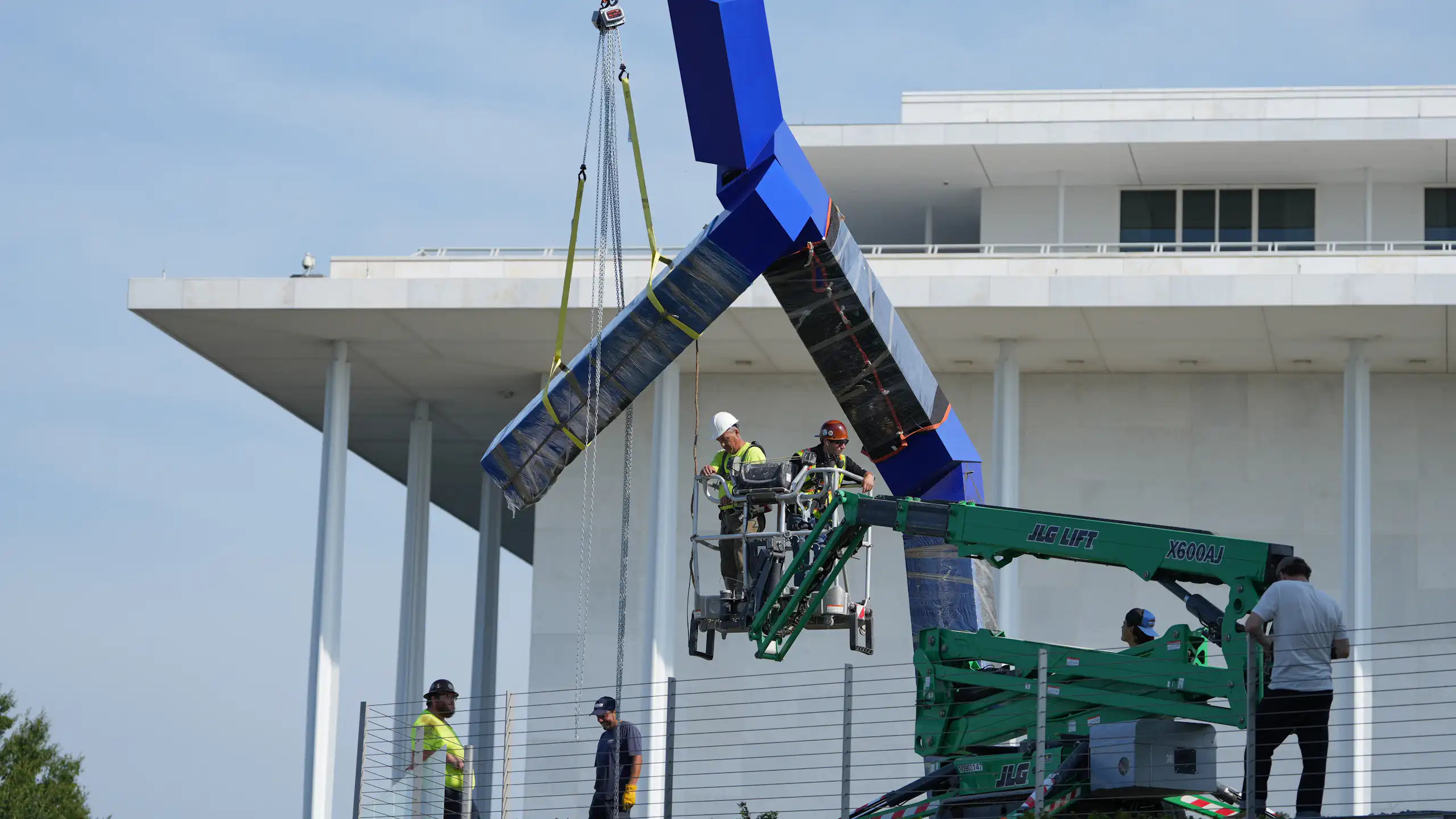 A crane with workers seen lifting a blue sculpture that seems to be in a large Y shape.