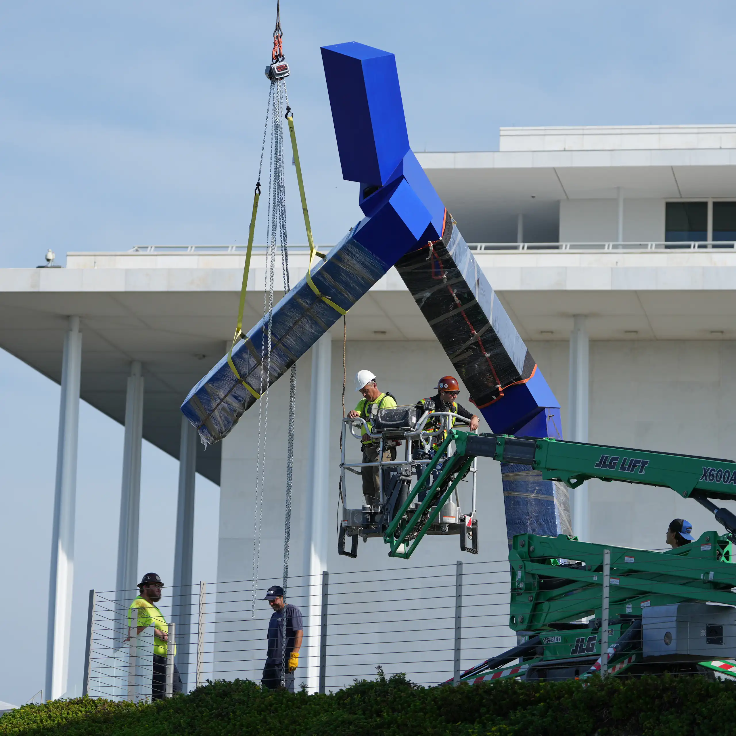 A crane with workers seen lifting a blue sculpture that seems to be in a large Y shape.