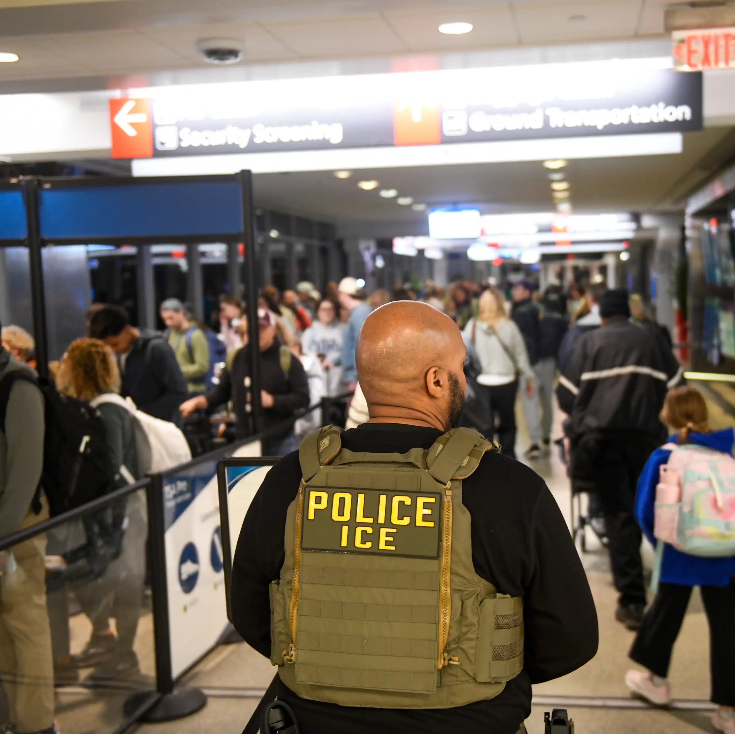 Man wearing green bullet-proof vest that says 'POLICE ICE' stands in busy area of airport