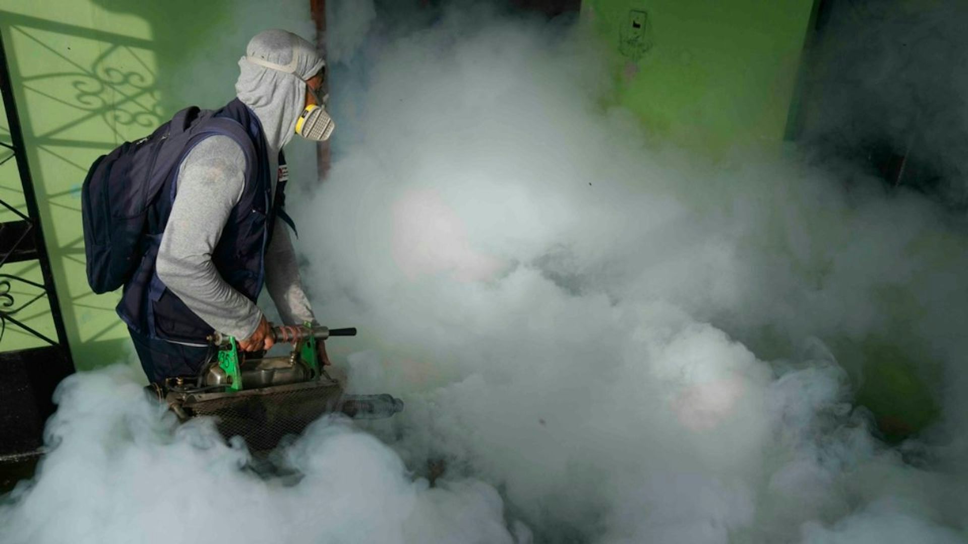 A health worker wearing white protective gear that covers from head to toe and a yellow mask fumigates for mosquitoes inside a home with a green wall.