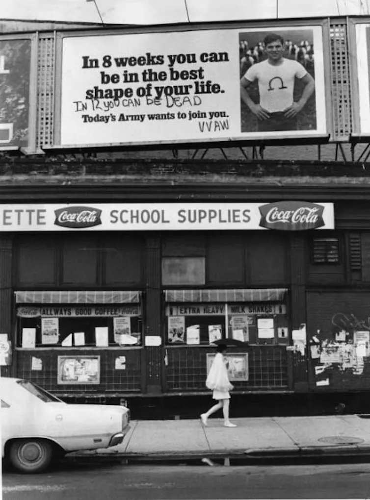 Black and white photo of street scene with billboard encouraging men to sign up for Army and anti-war graffiti on the billboard