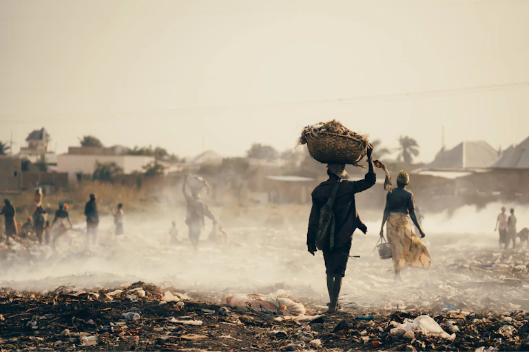 People walk across a flat garbage dump with smoke rising from the garbage