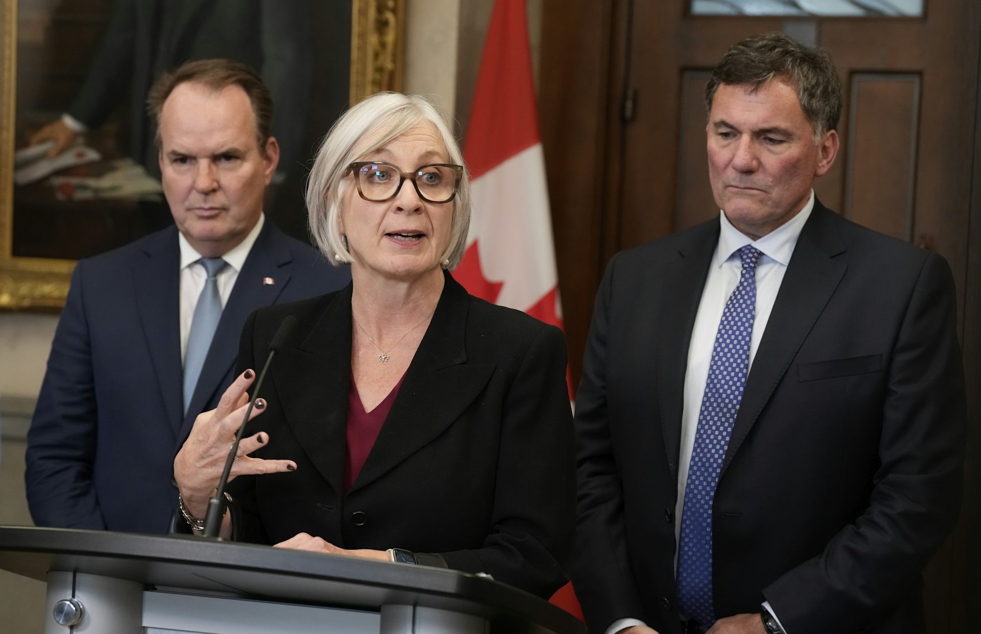 A white woman with a short bob in a blazer speaks from behind a podium, which two middle-aged white men in suits stand behind her