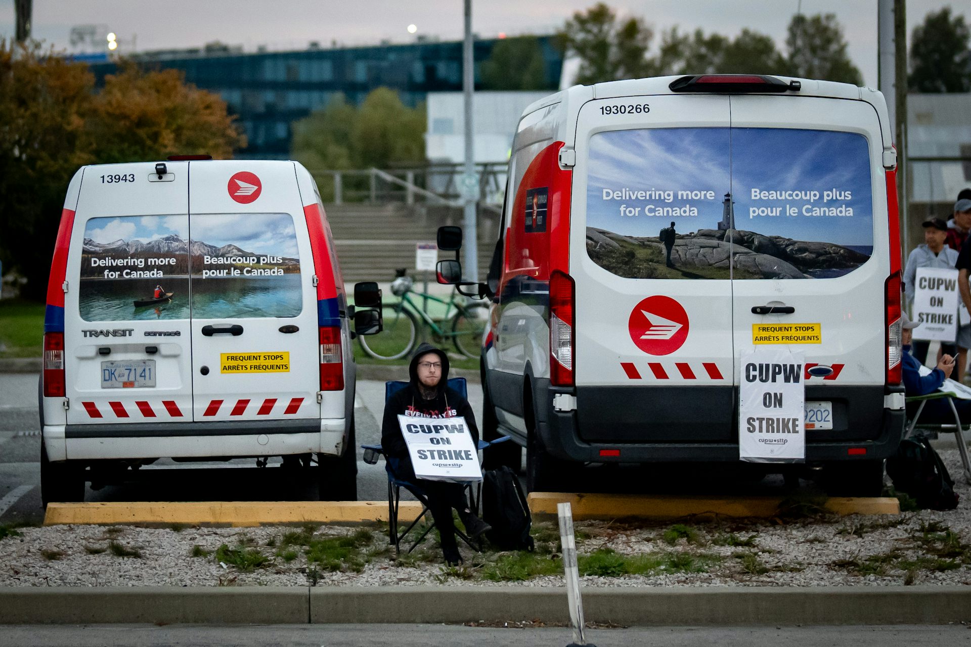 A person sits behind parked Canada Post vans, holding a sign that says 'CUPW on strike'