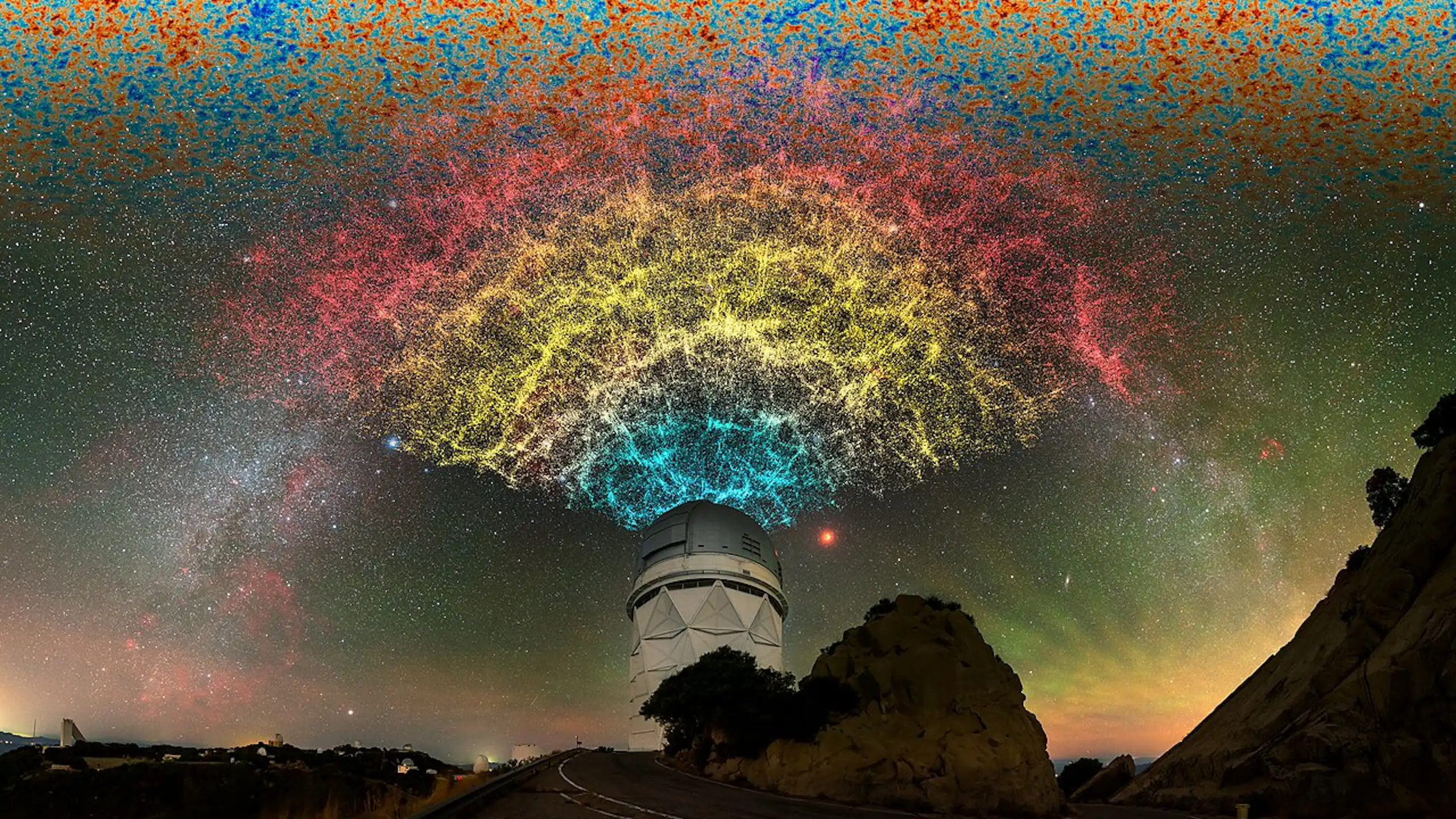 A dome telescope on top of a hill, the sky above illuminated with stars and yellow, blue and red light.
