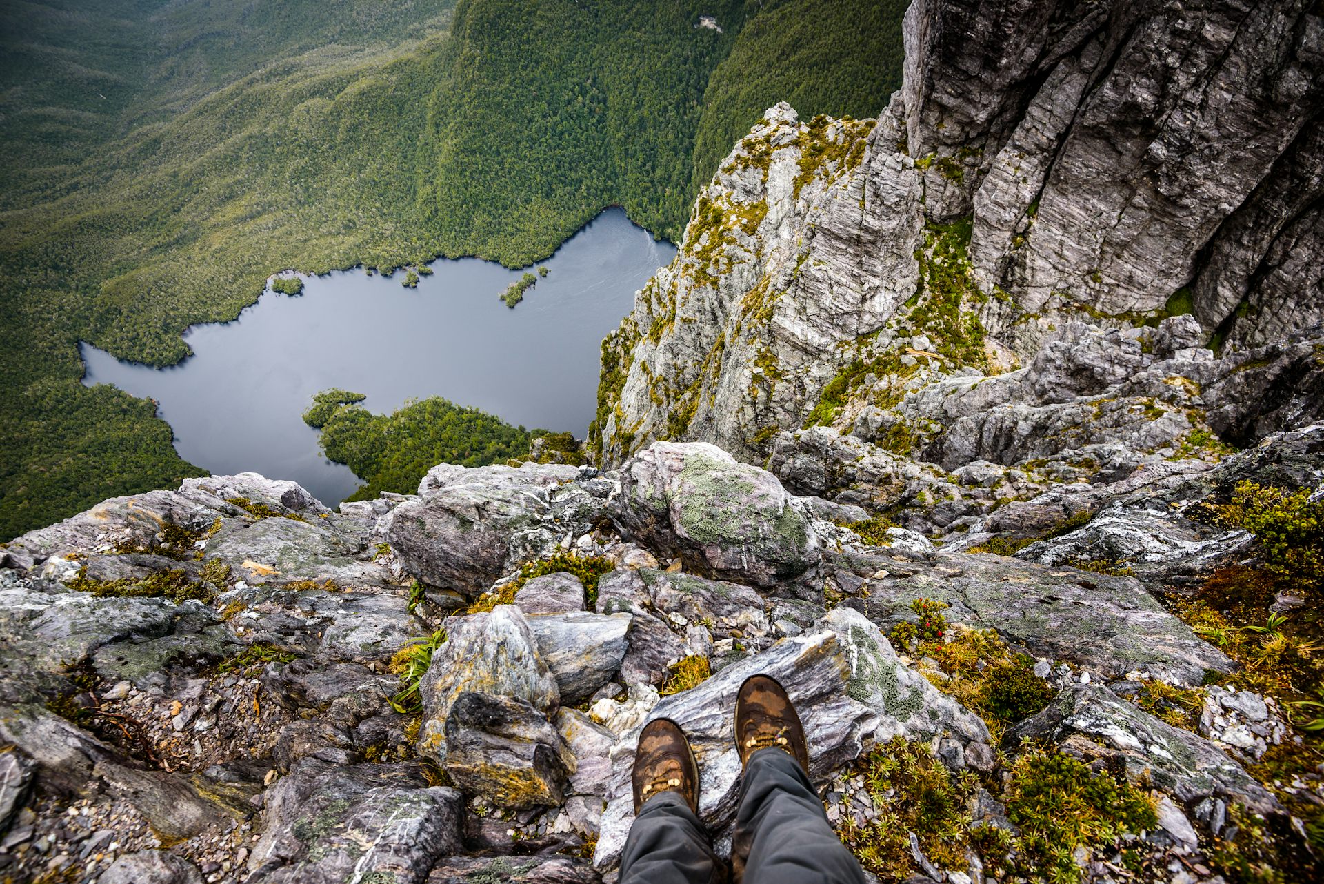 POV view of hiker's feet on rocks atop mountain, looking to lake far below.