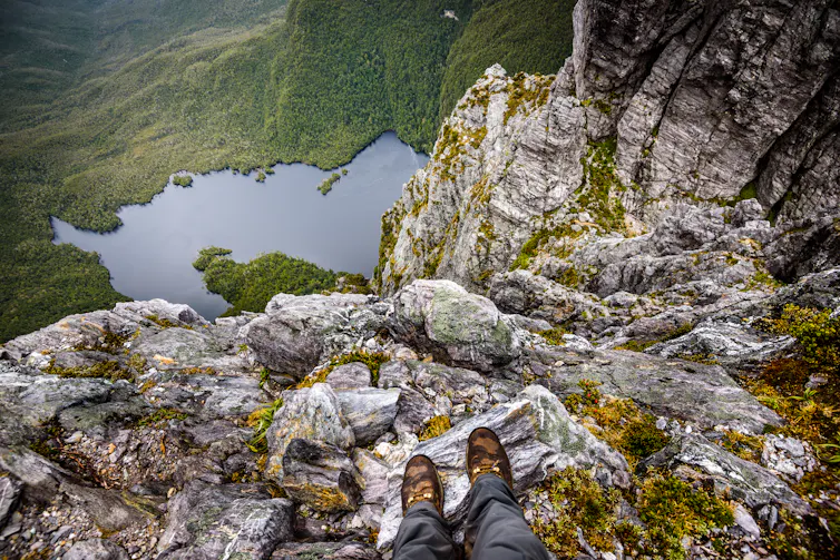 POV view of hiker's feet on rocks atop mountain, looking to lake far below.