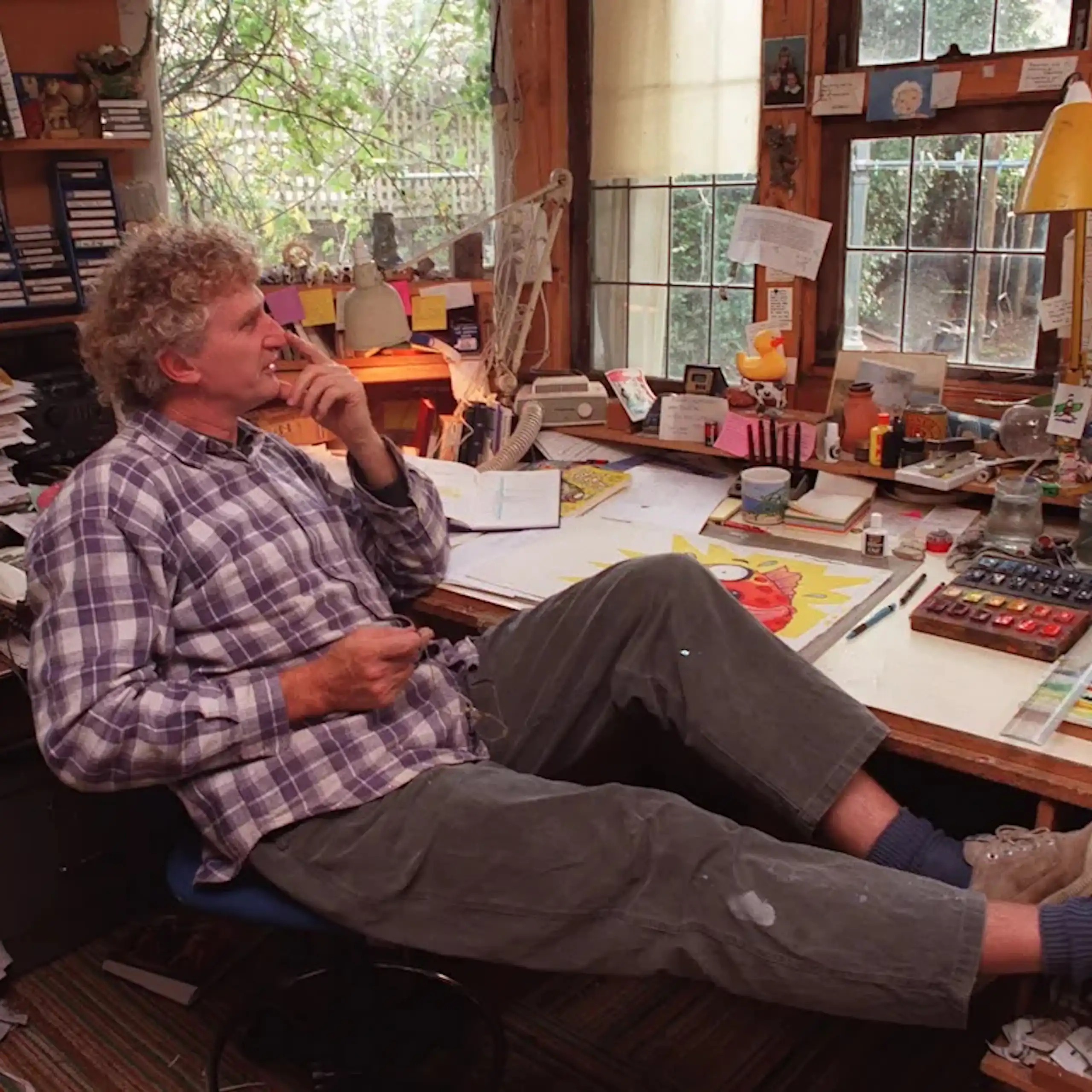 A man at a desk covered with papers and illustrations and art supplies, his feet up, dreaming out the window.