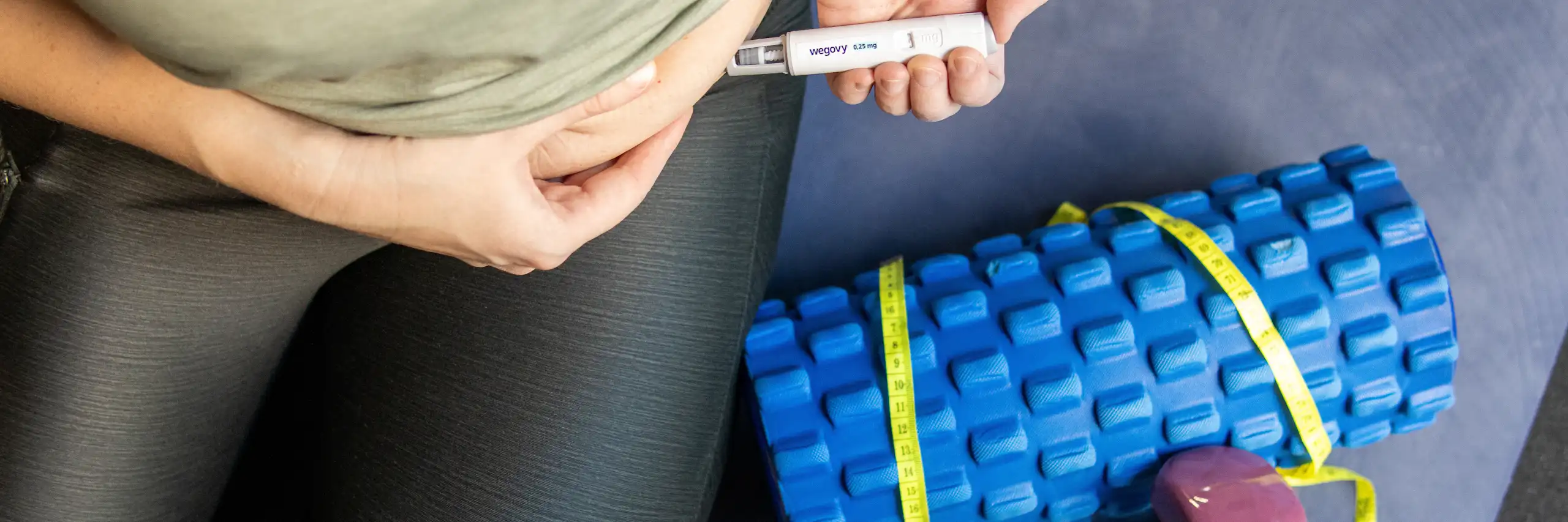 Une femme, en tenue de sport, en train de s’injecter une dose de médicament antiobésité Wegovy dans l’abdomen. Près d’elle, des haltères et un rouleau de mousse de gymnastique.