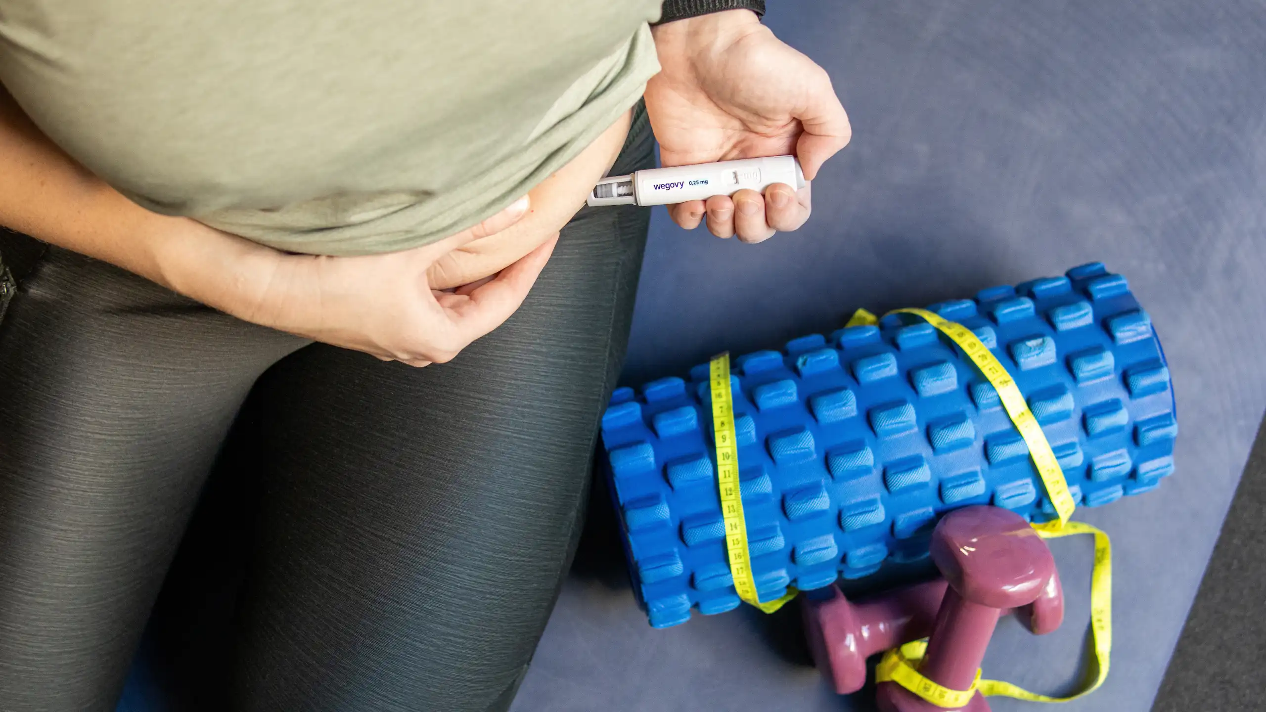 Une femme, en tenue de sport, en train de s’injecter une dose de médicament antiobésité Wegovy dans l’abdomen. Près d’elle, des haltères et un rouleau de mousse de gymnastique.