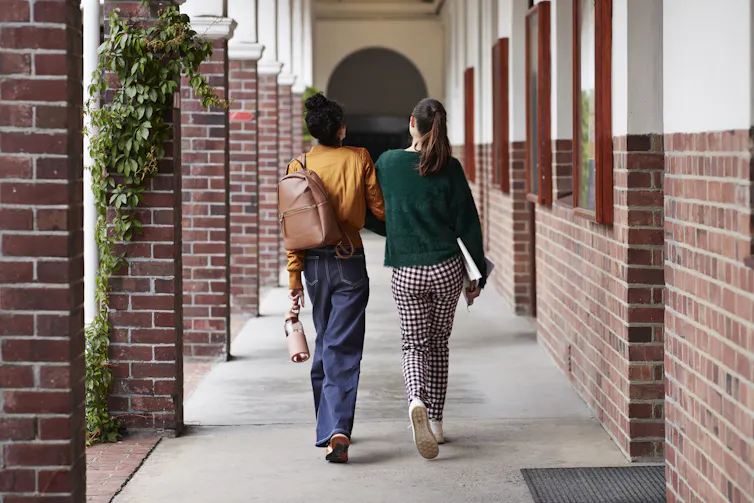 Two women walk closely together down a stone path with red brick pillars on either side of them.