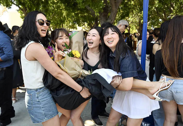 Four young women smile and pose as they lift one of the women, wearing a black graduation gown and carrying flowers, in the air.