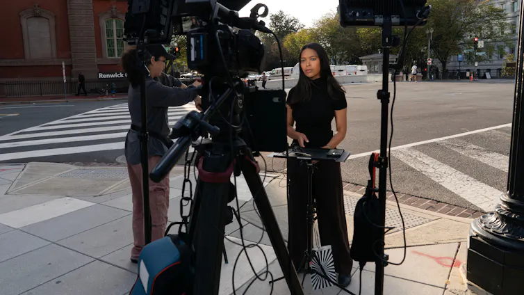 A woman stands on a street corner in front of a television news camera and other broadcast equipment.