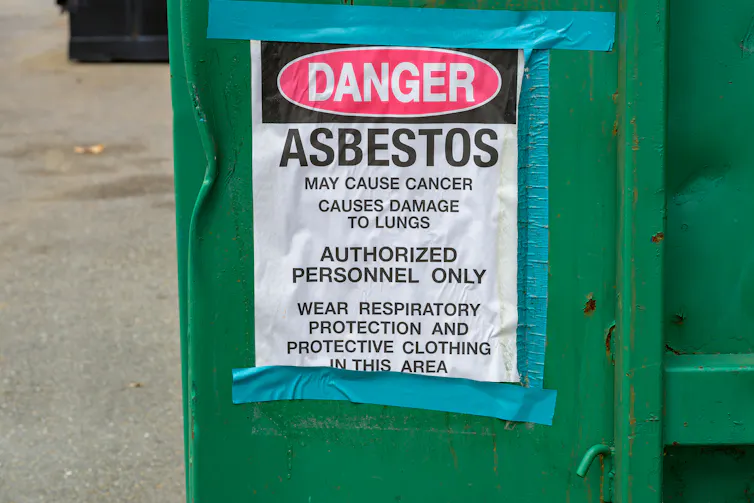 A sign explaining the danger of asbestos is taped to a dumpster.