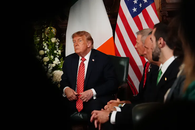A man in a suit sits flanked by two flags.