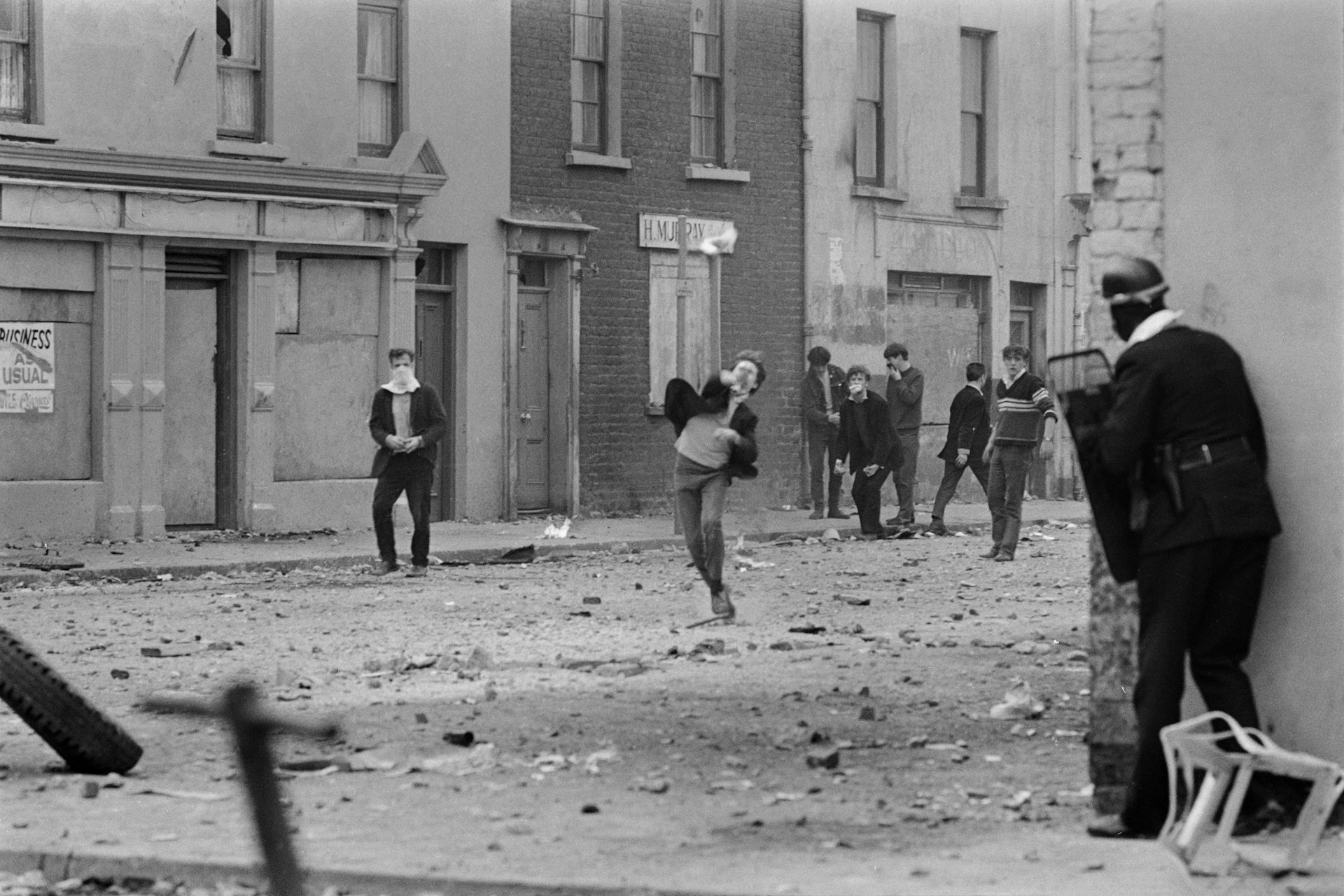 A youth throws a projectile toward a man in uniform carrying a shield.