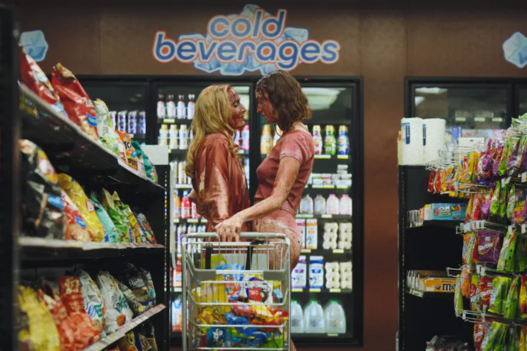 Two women stand close together, face to face, in a convenience store, hands intertwined on a shopping cart handle