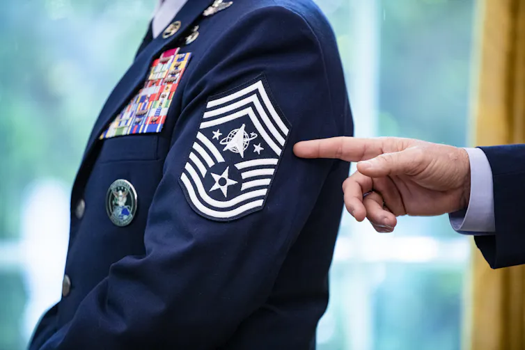 A hand points to a Space Force chevron rank insignia on the sleeve of a dark blue dress uniform
