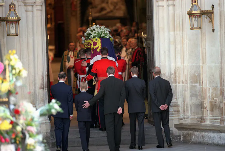 Three men and two boys walk behind a coffin in a funeral procession, one man with a hand on a boy's back to comfort him.