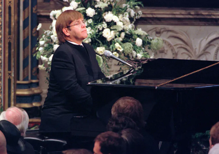 Elton John, dressed in black, sings at Diana's funeral, white roses in background.