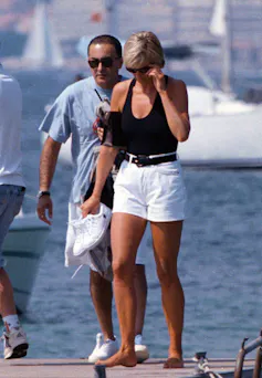 A beautiful woman, Diana, with white short shorts and black bathing suit, walking, head down, with a man behind her.