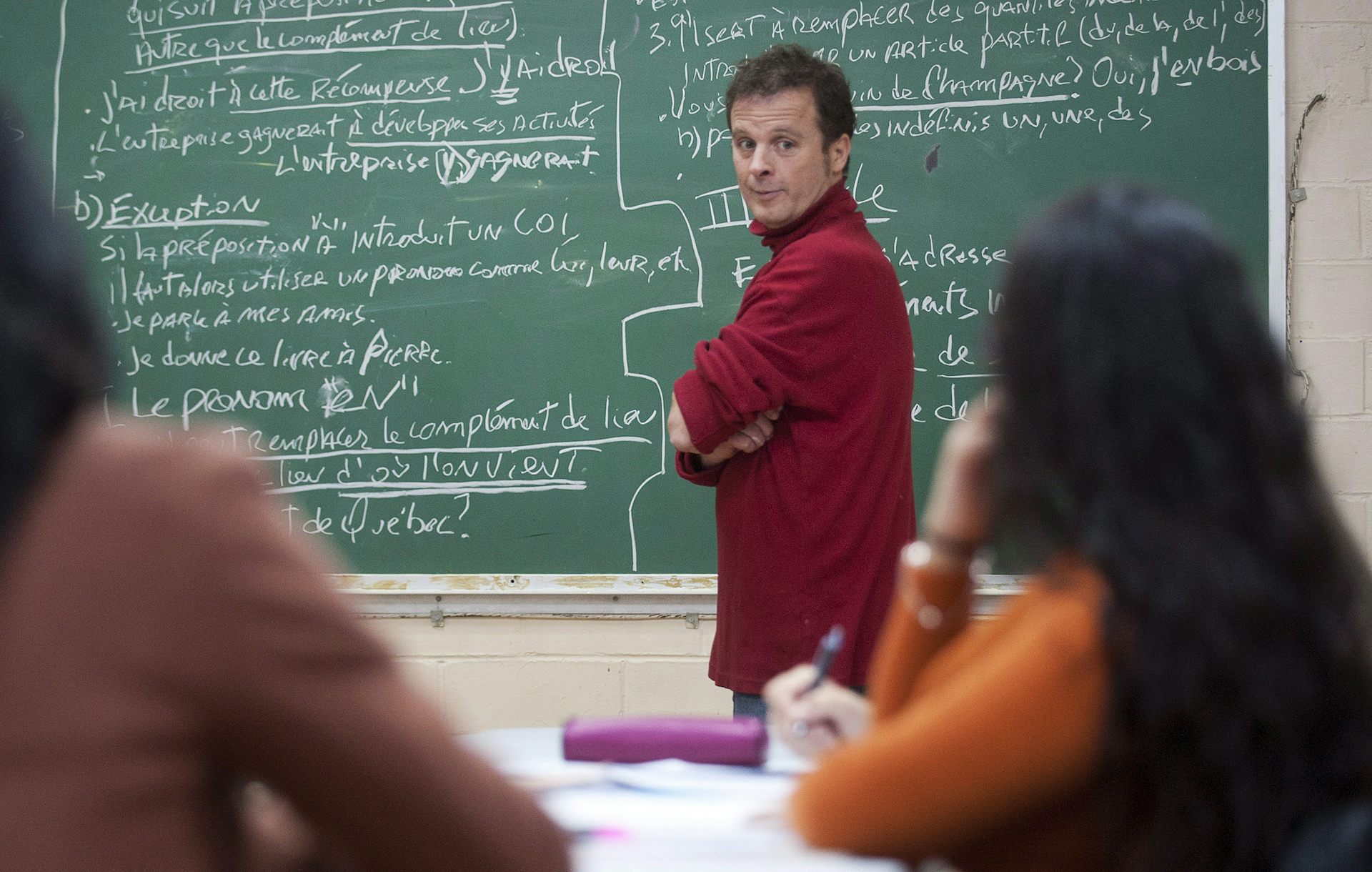 A middle-aged white man stands in front of a chalkboard at the front of a classroom
